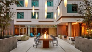 A modern outdoor dining area with a long wooden table surrounded by chairs, a fire feature in the center, and hanging pendant lights.