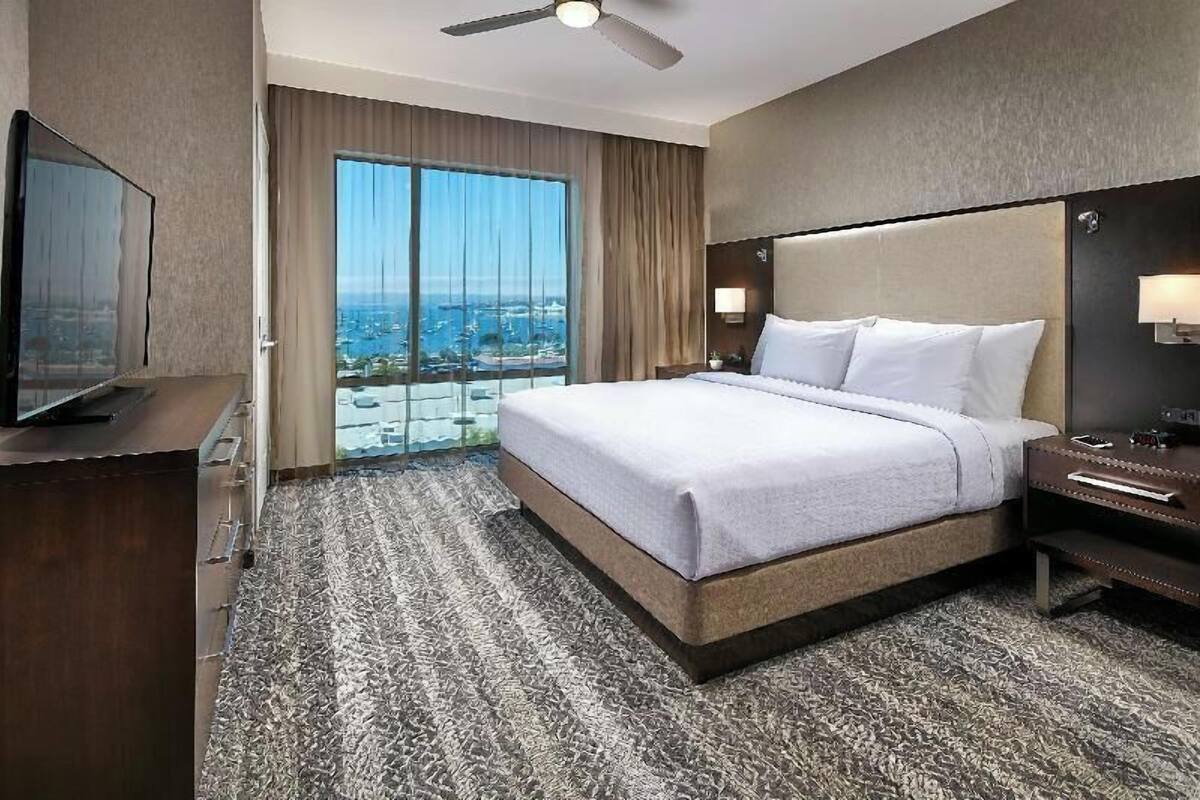 A hotel room with a large bed, a television, a nightstand, and a view of the city through the window.