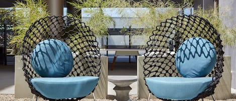 Two chairs with blue cushions and a small table in between, set on a patio with plants and a building in the background.