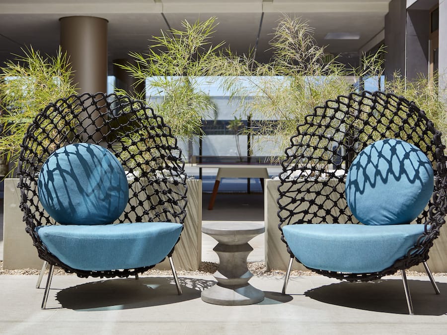 Two chairs with blue cushions and a small table in between, set on a patio with plants and a building in the background.
