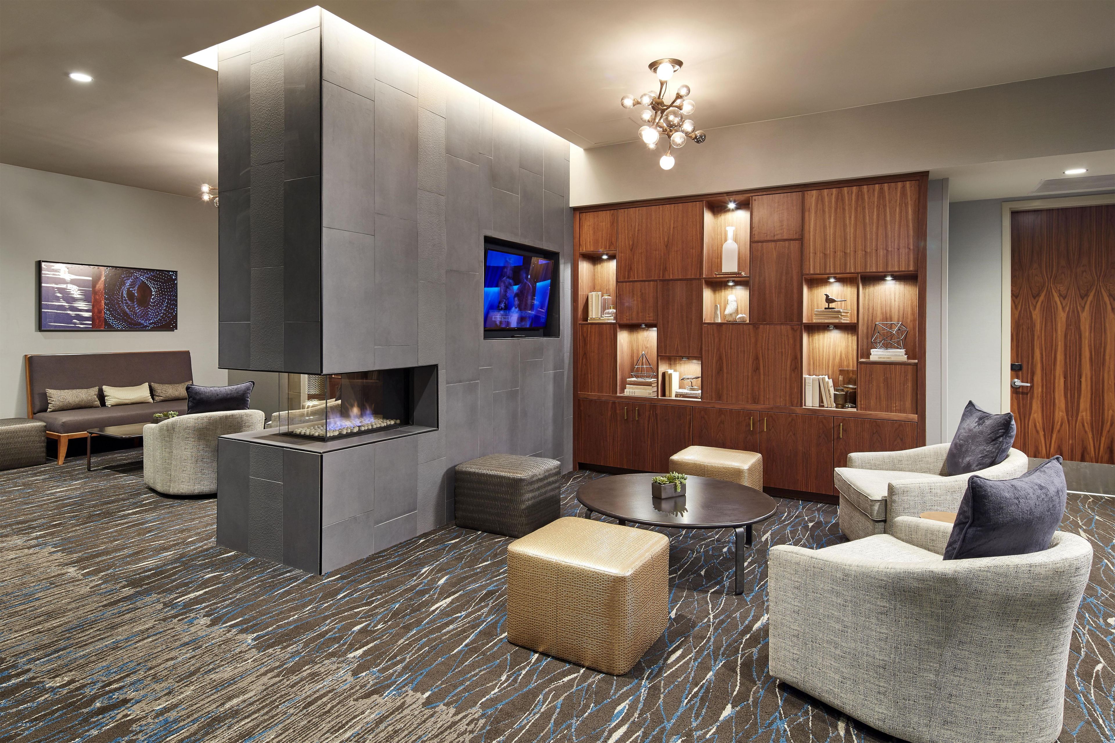 A modern hotel lobby with a fireplace, comfortable seating, a flat-screen TV, and a wooden display cabinet.