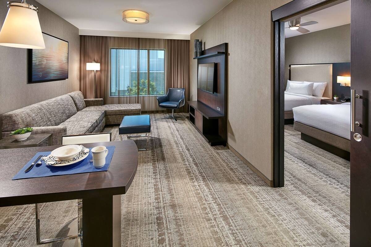 A hotel room with a sofa, a TV, a bed, a dining table with a plate and cup, and a window with curtains.