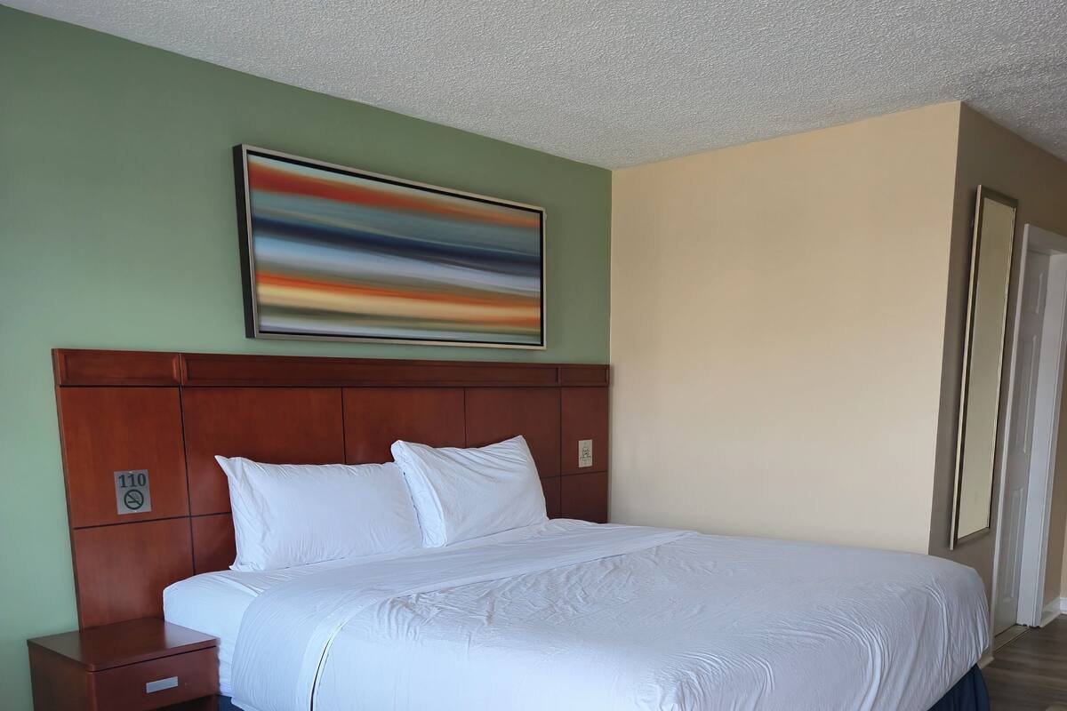 Ein Hotelzimmer mit einem großen Bett, einem Nachttisch aus Holz, einem Gemälde an der Wand und einer Tür auf der rechten Seite.