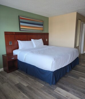 Habitación de hotel con una cama grande, una mesita de noche de madera, un cuadro en la pared y una puerta a la derecha.