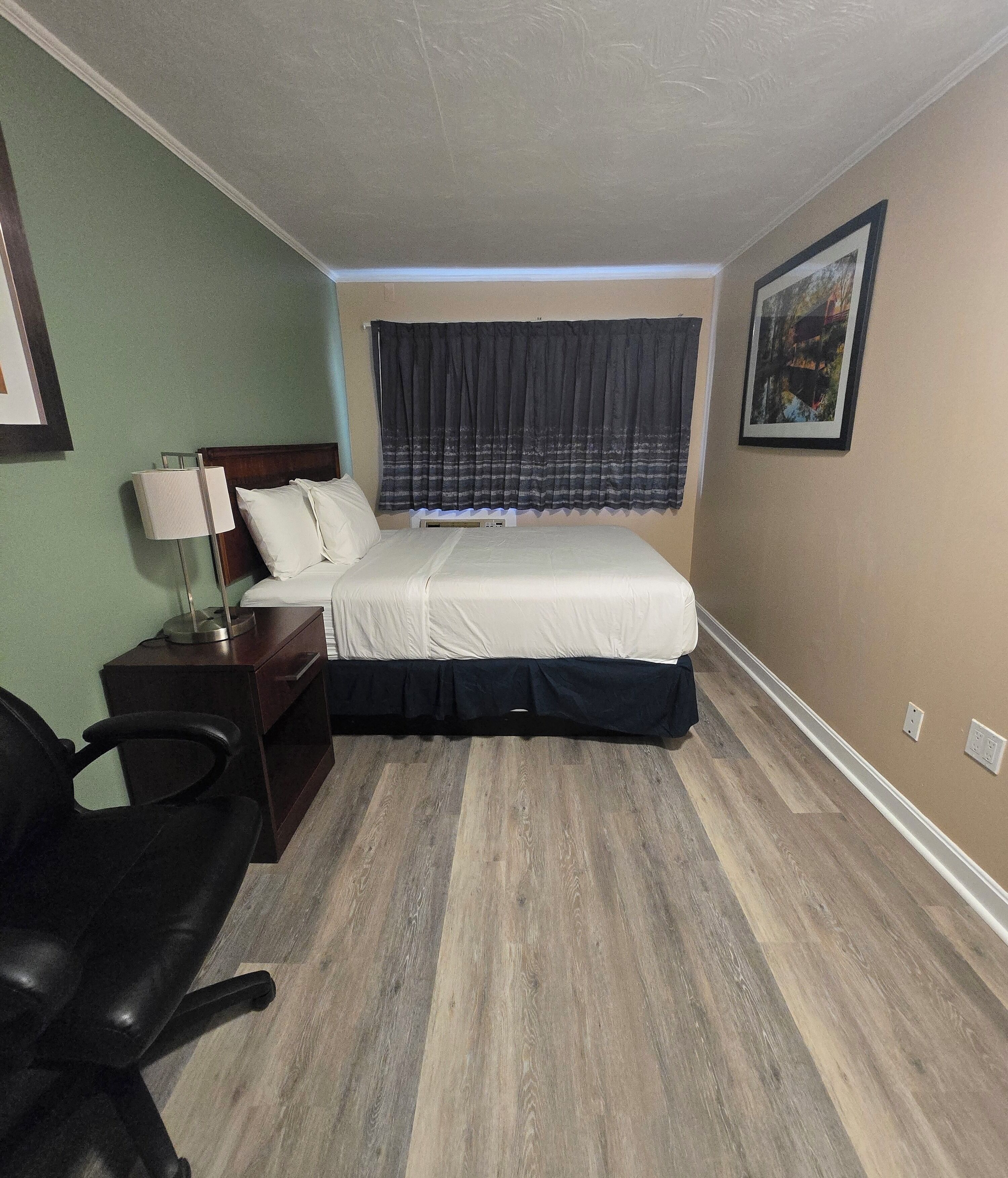 A small, neatly arranged hotel room with a bed, bedside table, lamp, chair, and a window with curtains.