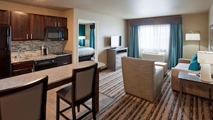 A hotel room with a kitchenette, a bed, a seating area with a sofa and chairs, a TV, and a window with curtains. - GrandStay Hotel and Suites (Valley City)