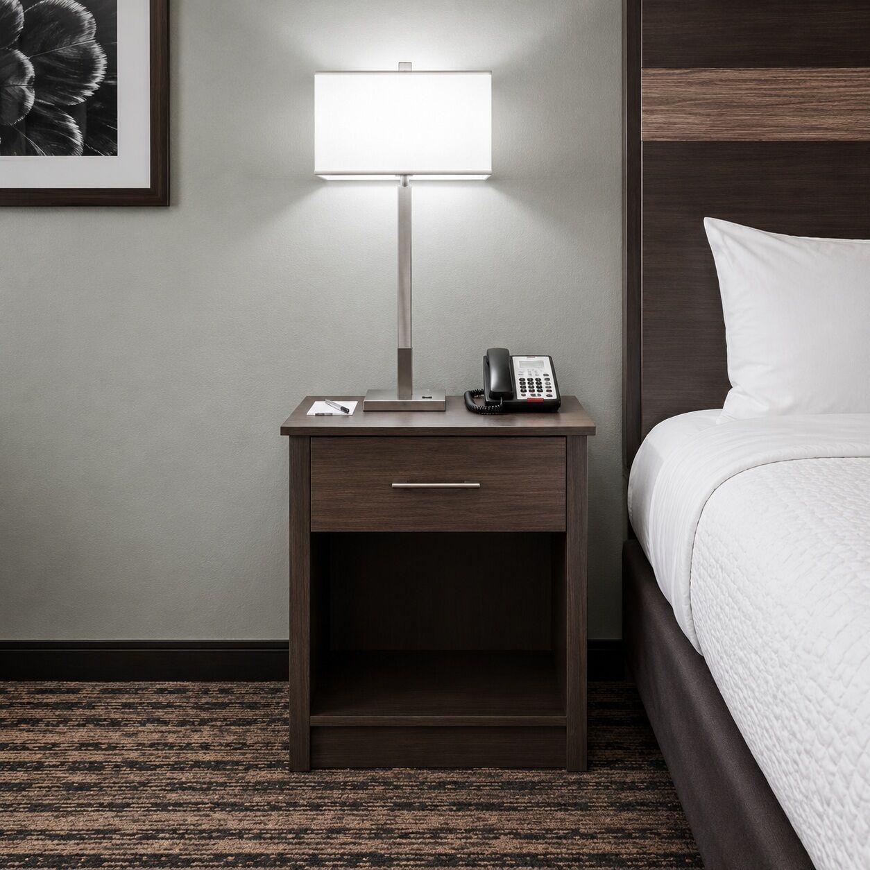 Habitación de hotel con mesita de noche, lámpara, teléfono y cama con edredón blanco.