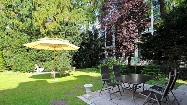 A patio with a table and chairs, an umbrella, and a statue in a garden setting. - Hotel Leopold (Munich)