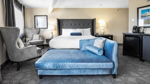 A hotel room with a large bed, a grey armchair, a blue sofa, a desk with a lamp, and a small refrigerator. - Aqua Blue Hotel (Narragansett)