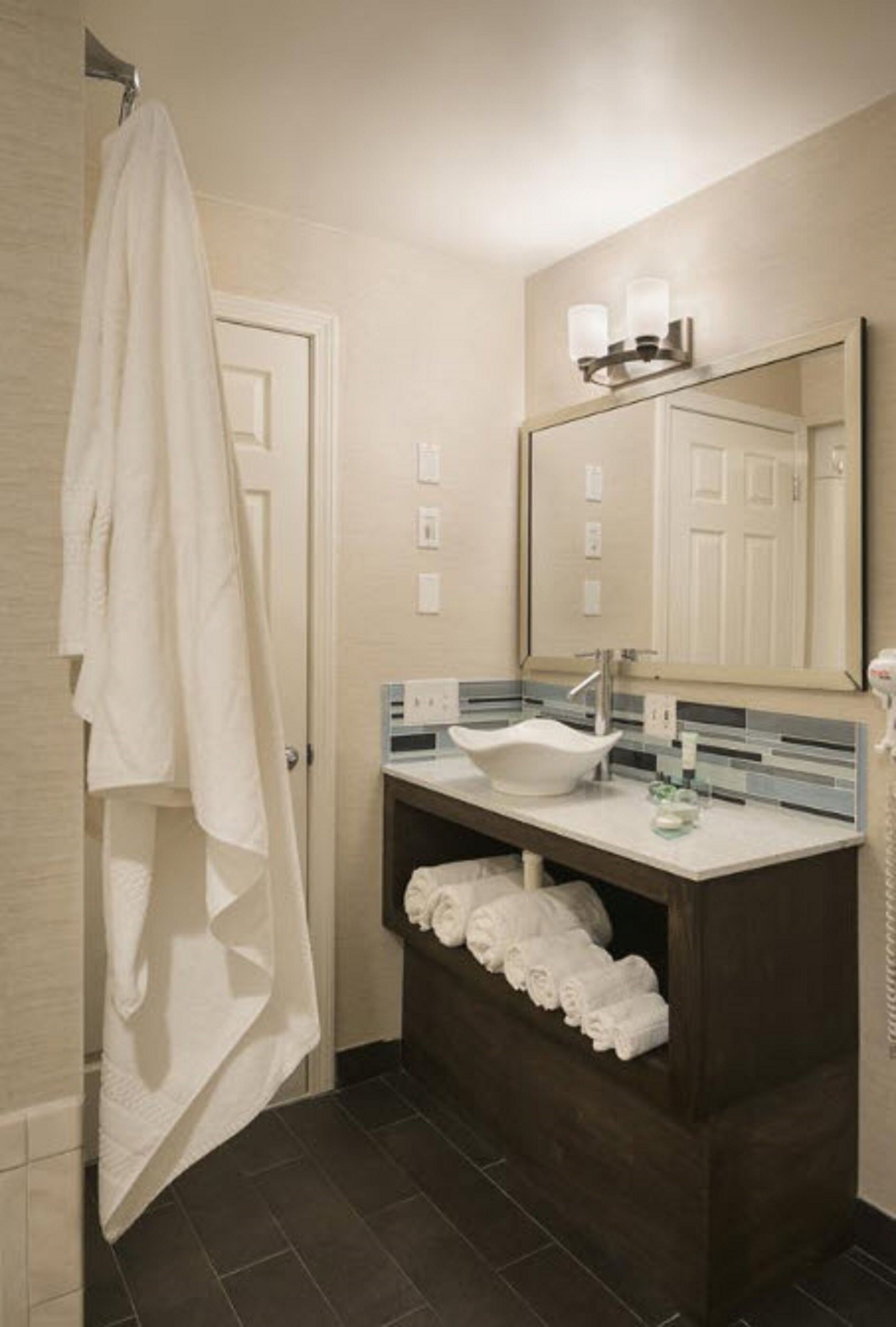Un baño con lavamanos blanco, mueble de tocador de madera oscura, un espejo grande y un perchero para toallas.