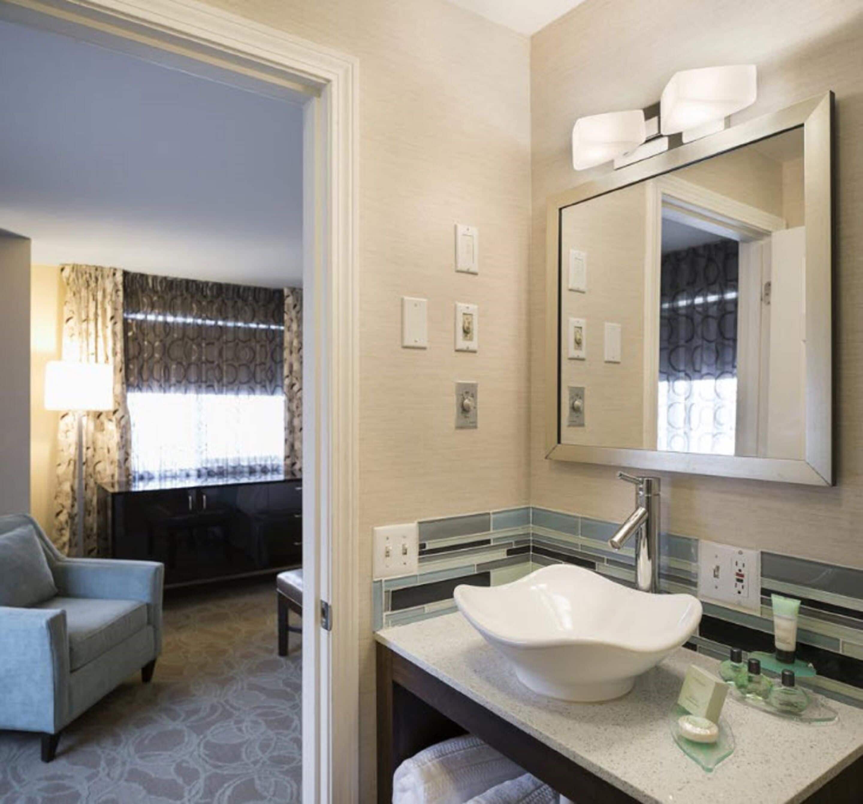 Un baño de hotel con un lavamanos grande, un espejo y una silla en la habitación contigua.