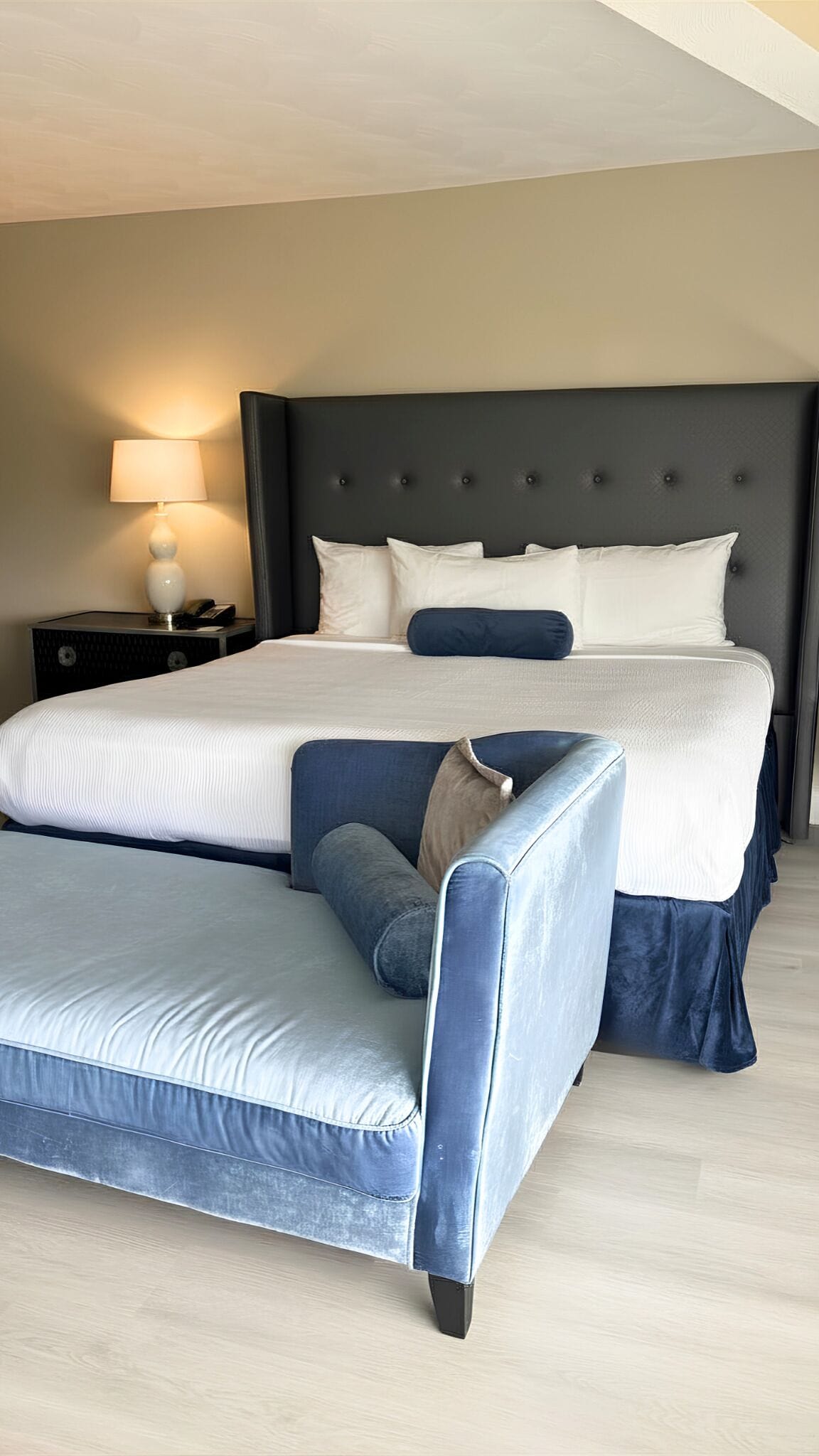 Un lit avec une tête de lit sombre, une table de chevet avec une lampe et un banc recouvert d’un tissu bleu.