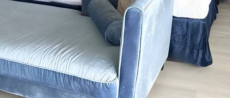 Un lit avec une tête de lit sombre, une table de chevet avec une lampe et un banc recouvert d’un tissu bleu.