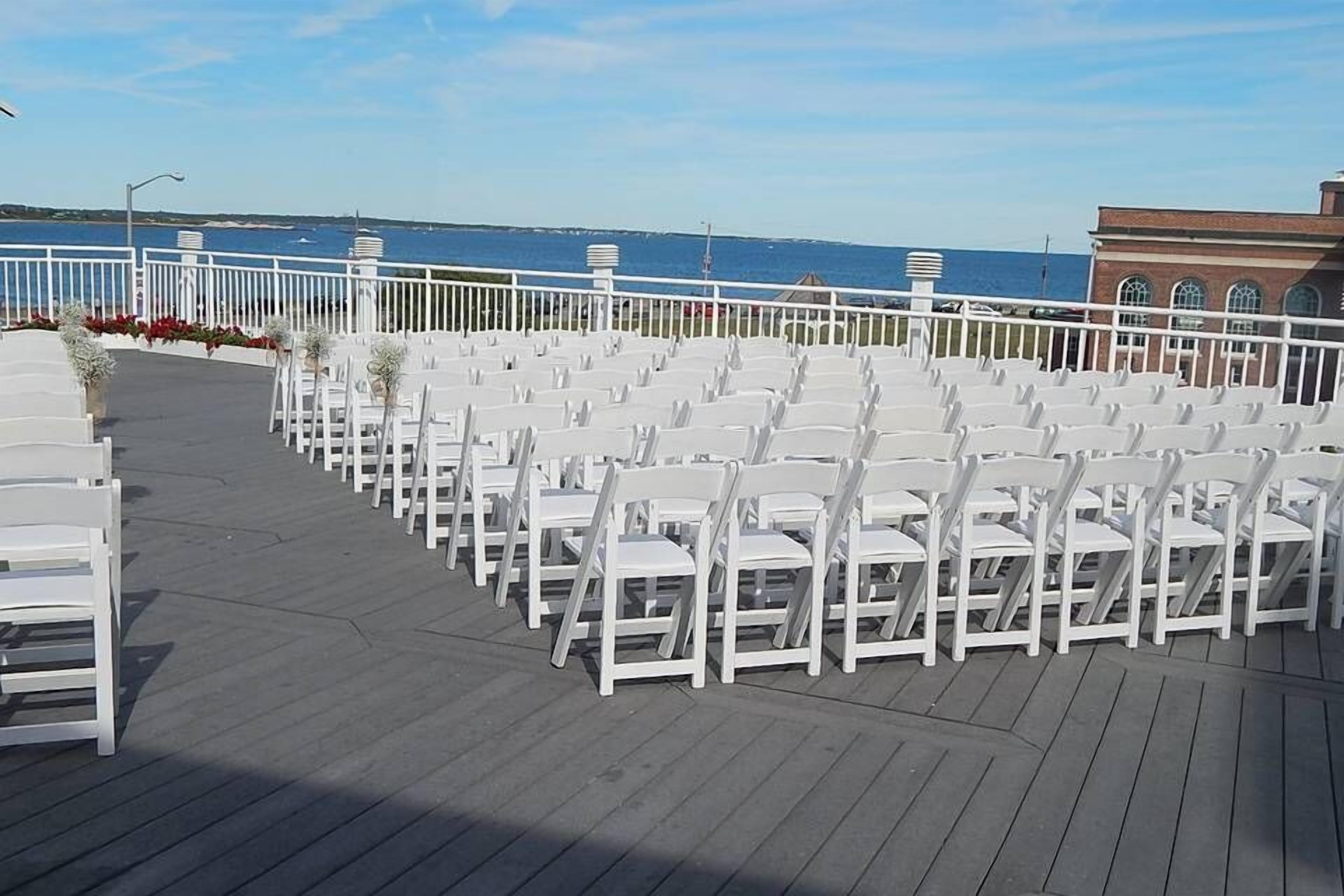 An outdoor seating area with white chairs arranged in rows, a wooden deck, and a view of the sea.
