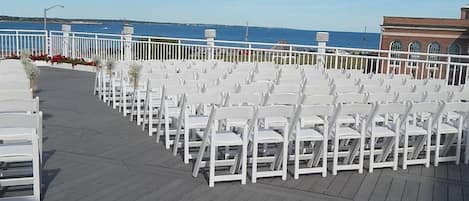 Un área de estar al aire libre con sillas blancas dispuestas en filas, una terraza de madera y vistas al mar.