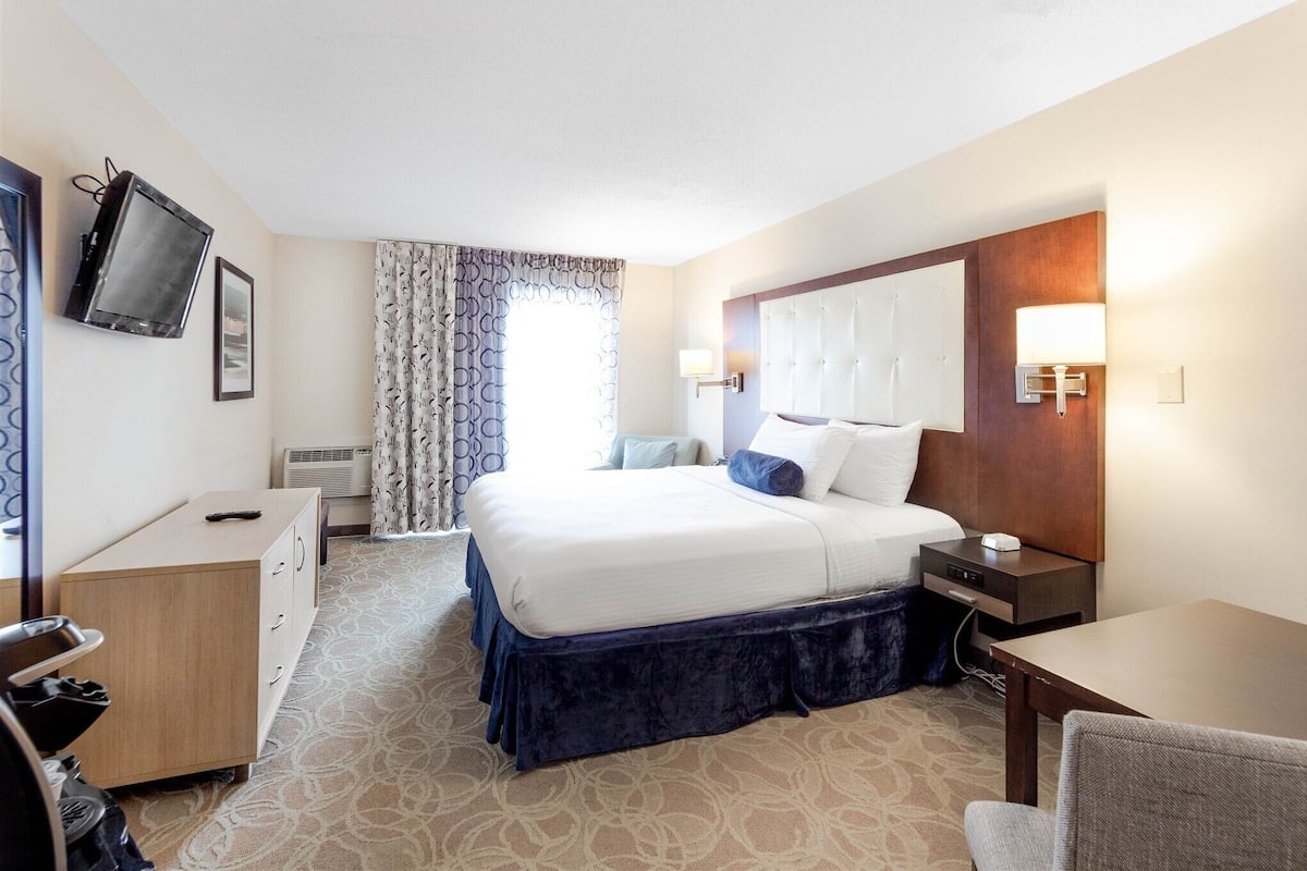 A hotel room with a bed, bedside table, lamp, TV, and a desk with a chair.