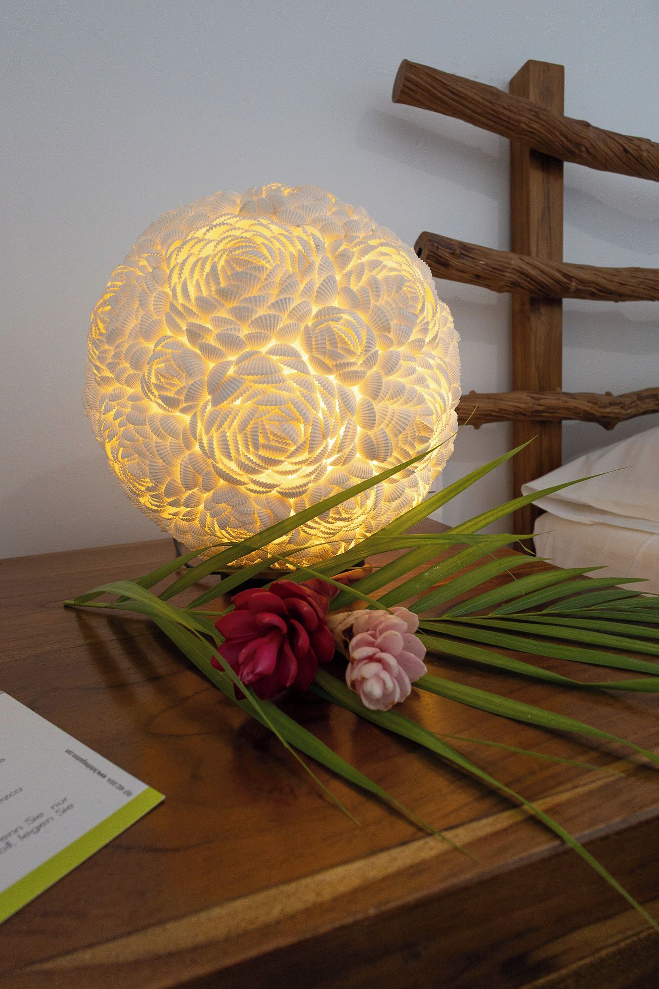 A decorative light fixture with a textured surface, placed on a wooden table with green leaves and red flowers.