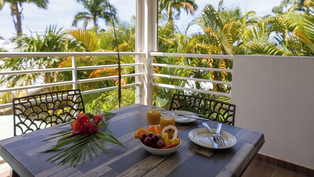 A table with a fruit bowl, a glass of orange juice, and a plate with a fork on a balcony with a view of palm trees. - Majesty Palm (Saint-François)