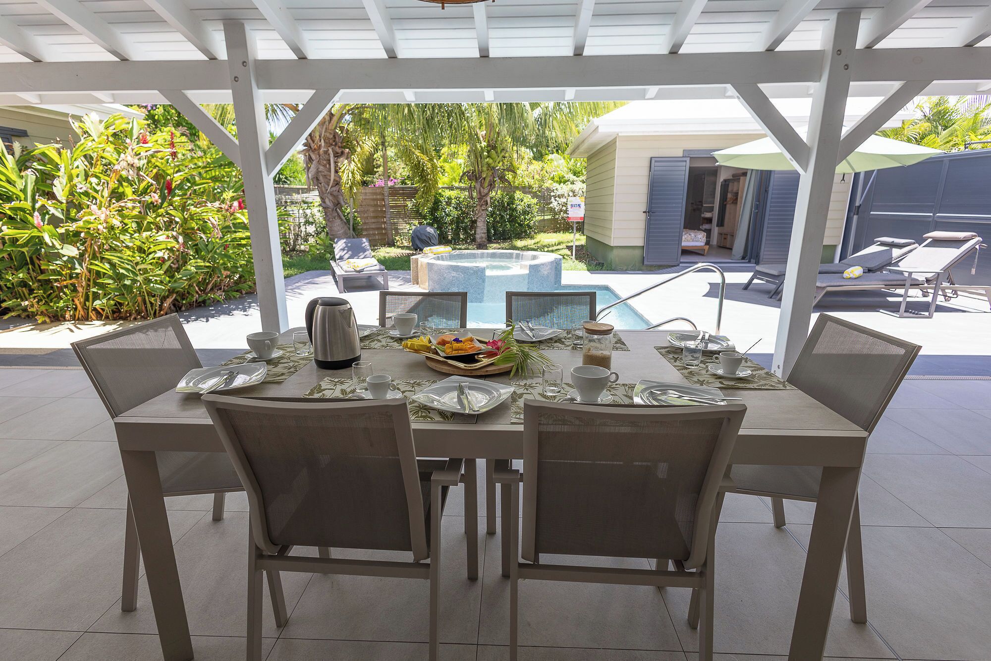A covered outdoor dining area with a table set for two, chairs, and a view of a swimming pool and garden.