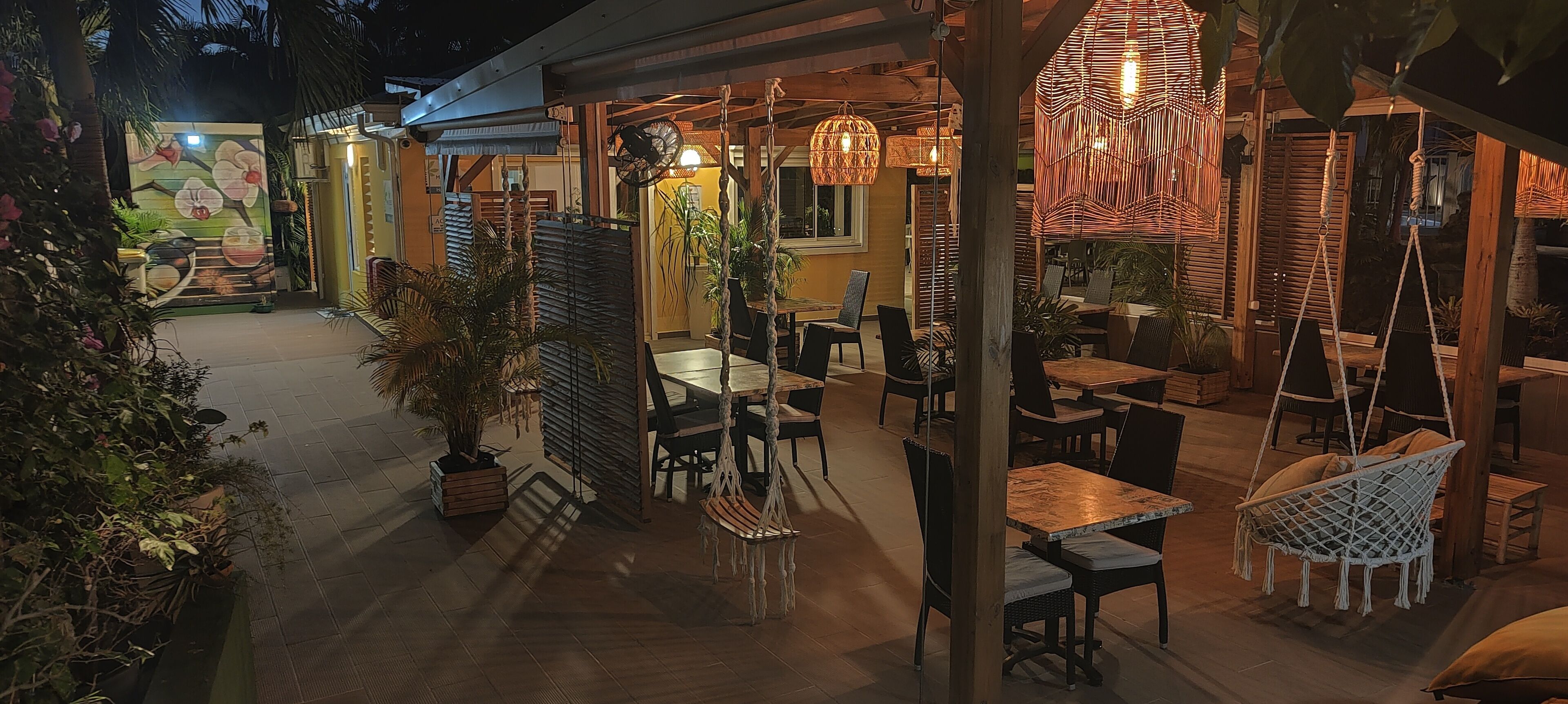 Un espace de restauration en plein air avec des tables et des chaises en bois, des fauteuils suspendus en rotin et des plantes en pot.