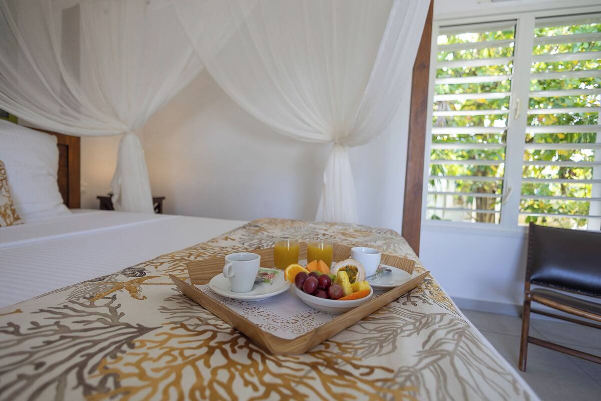 Ein Bett mit Baldachin, ein Tablett mit Frühstück, darunter Saft, Obst und eine Tasse, sowie ein Fenster mit Blick auf Grünflächen.
