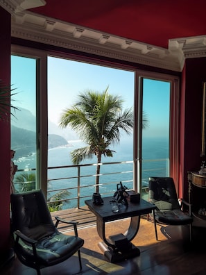 A room with a large window overlooking a coastal view, a palm tree, and two chairs.