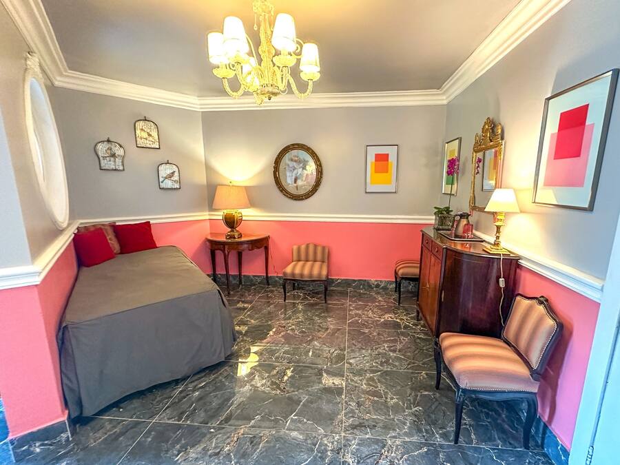 Une chambre d’hôtel avec un lit, deux chaises, un bureau, une lampe, un lustre et des œuvres d’art accrochées aux murs.