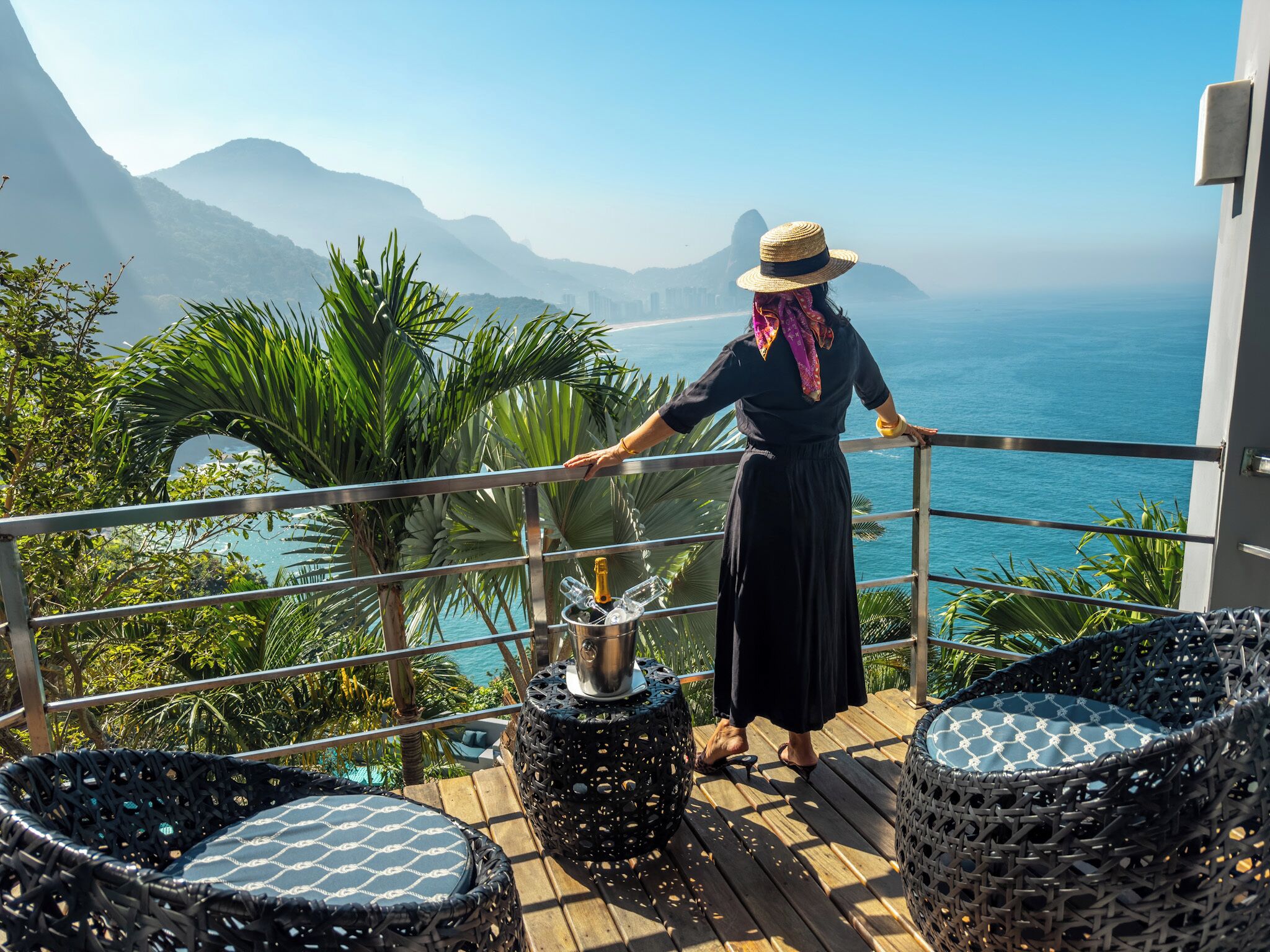 A balcony with a view of the ocean, a person standing by the railing, and two wicker chairs.