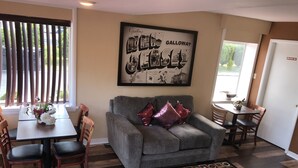 A living room with a grey sofa, a dining table with chairs, a wooden floor, and a framed picture on the wall.