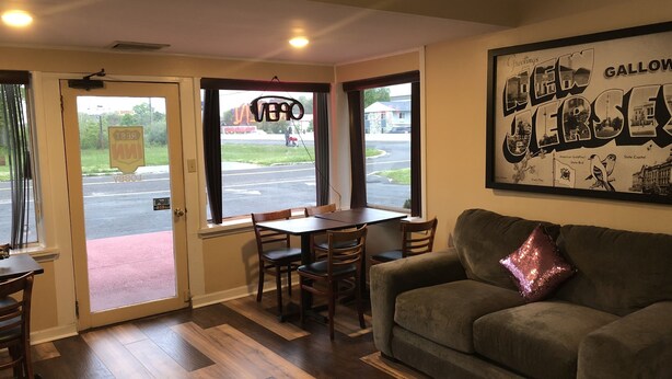 A room with a wooden floor, a green couch, a table with chairs, and a window with a view of a street and a sign that reads "NEVER".