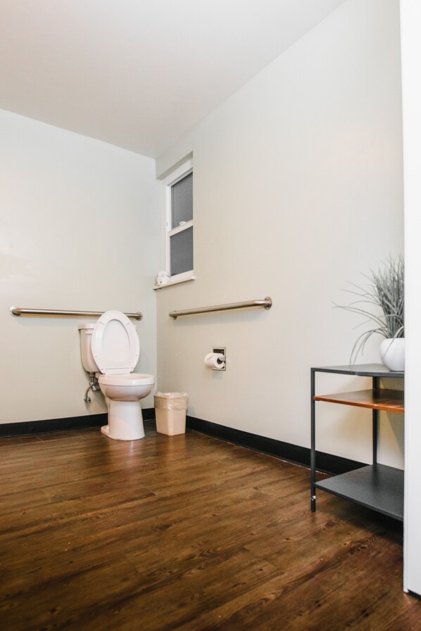 A bathroom with a toilet, wooden flooring, a handrail, a small window, and a plant on a shelf.