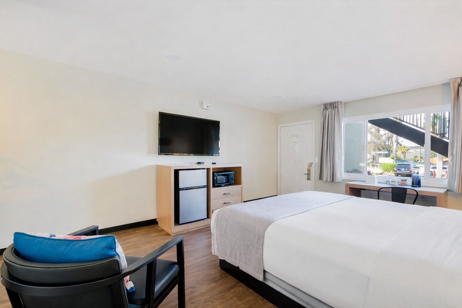 Habitación de hotel con una cama, un escritorio con computadora, televisor, refrigerador pequeño y ventana con vista al exterior.