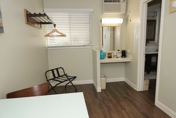 A small, well-lit room with a wooden chair, a table, a hanging coat rack, a mirror, and a shelf with toiletries.