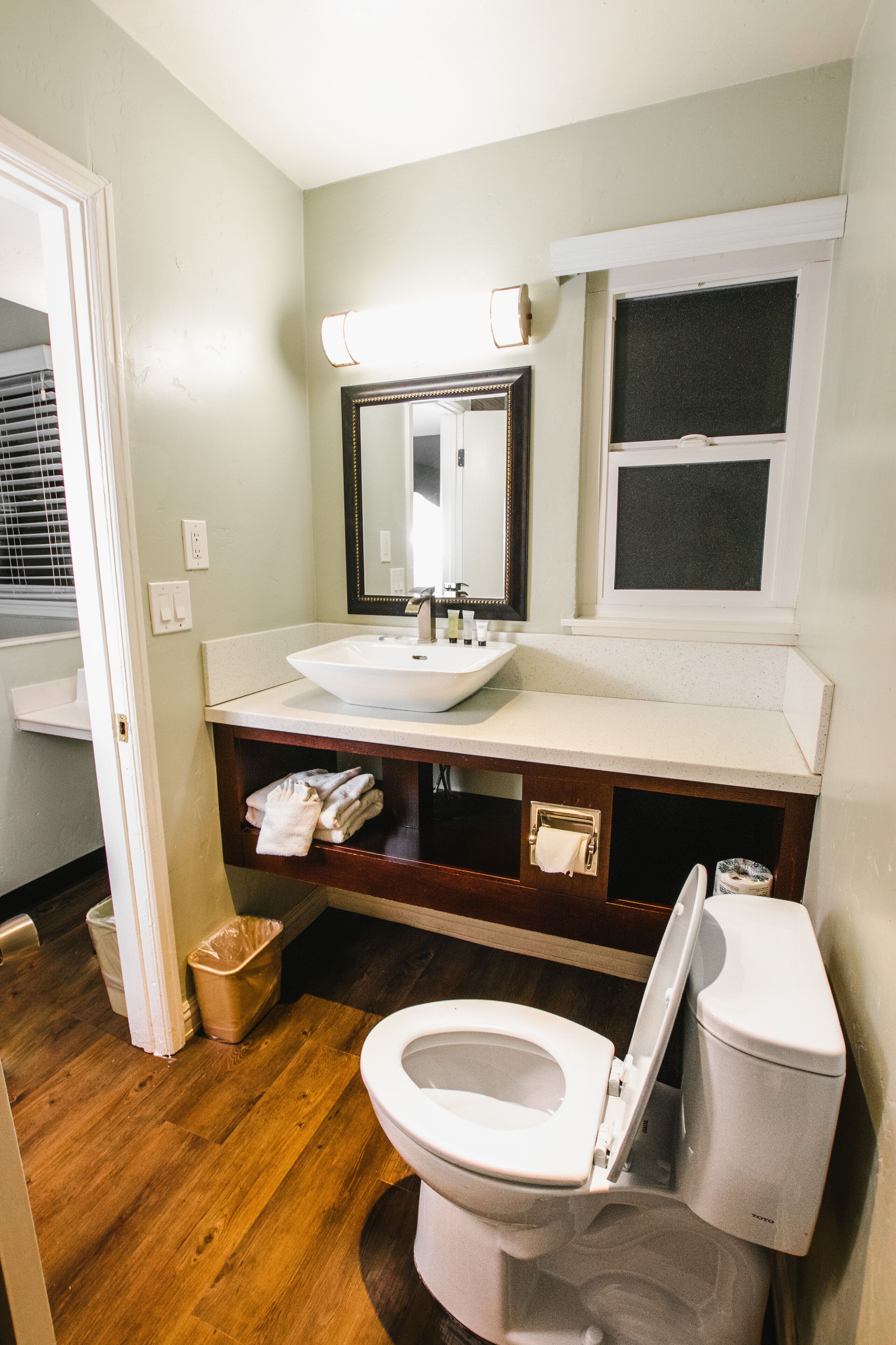 A bathroom with a white sink, mirror, and toilet. There is a towel rack with towels and a trash can.