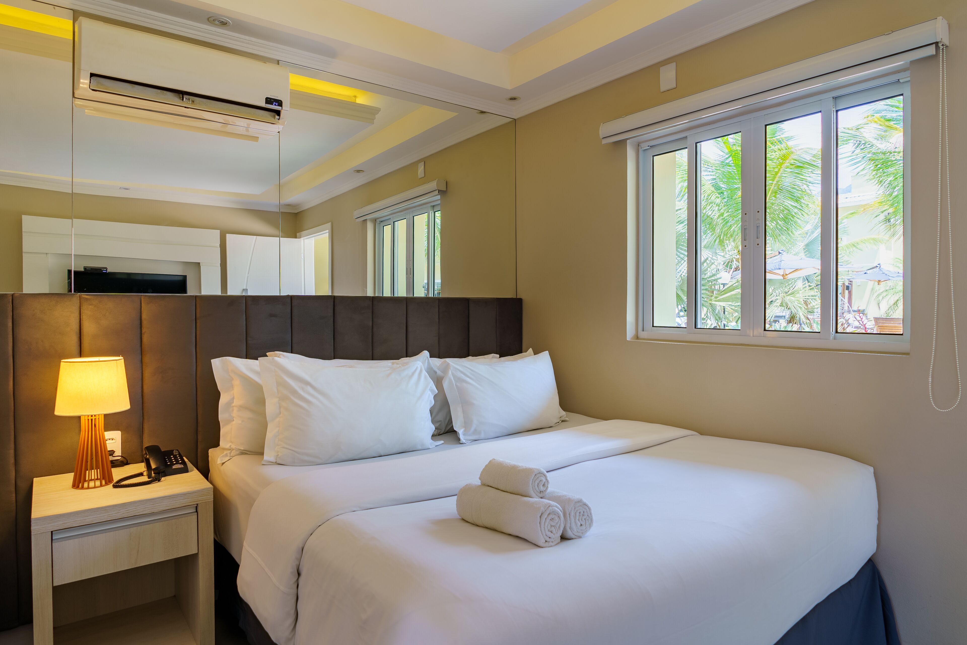 Habitación de hotel con cama, mesita de noche con lámpara, teléfono y ventana con vistas a palmeras.