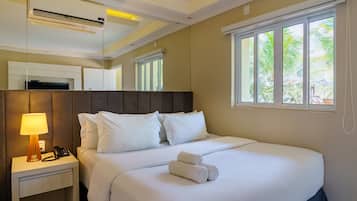 Habitación de hotel con cama, mesita de noche con lámpara, teléfono y ventana con vistas a palmeras.