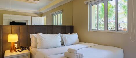 A hotel room with a bed, bedside table with a lamp, a telephone, and a window with a view of palm trees.