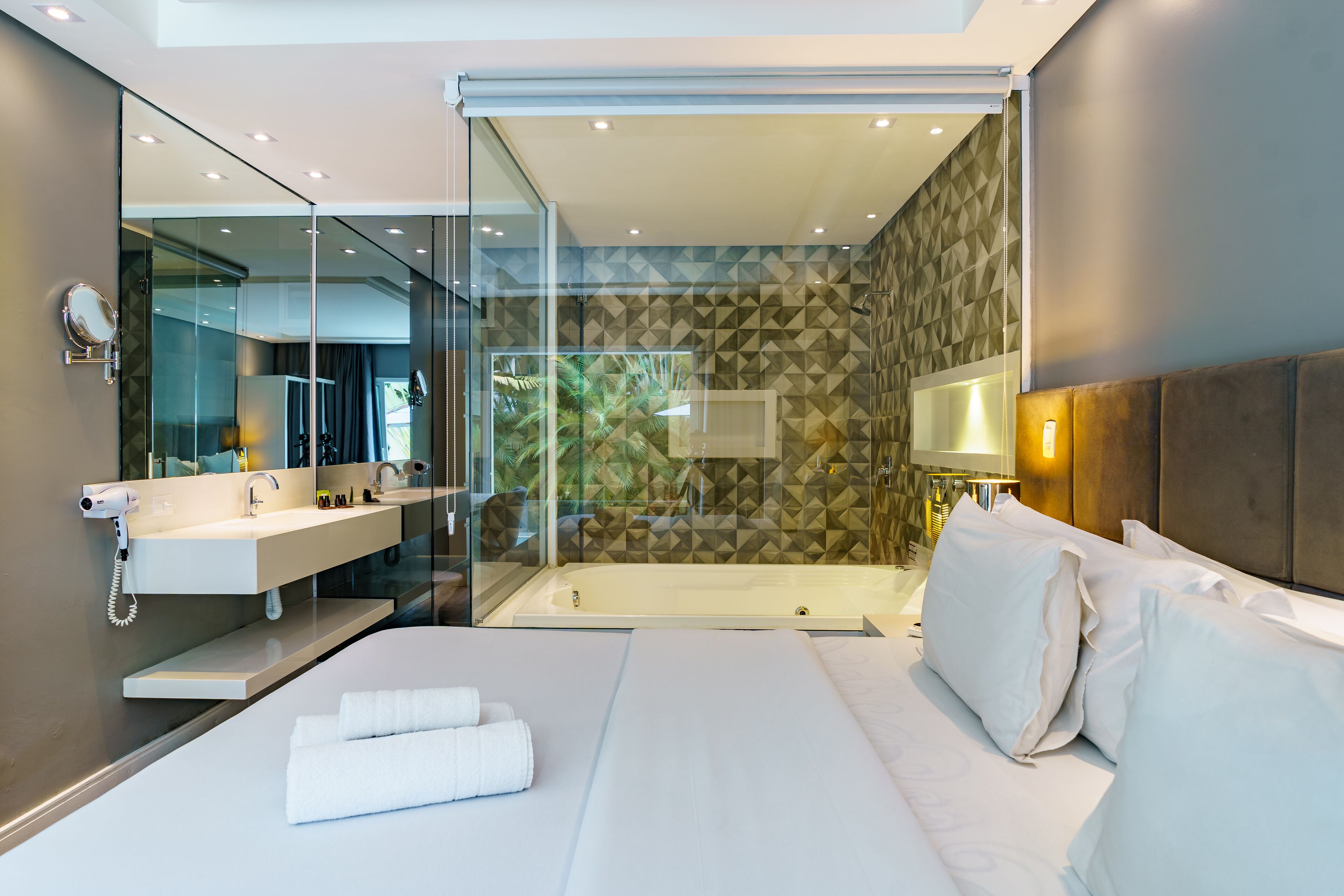 A modern hotel room with a glass-enclosed shower, a large bed with white linens, a vanity with a mirror, and a geometric-patterned wall.