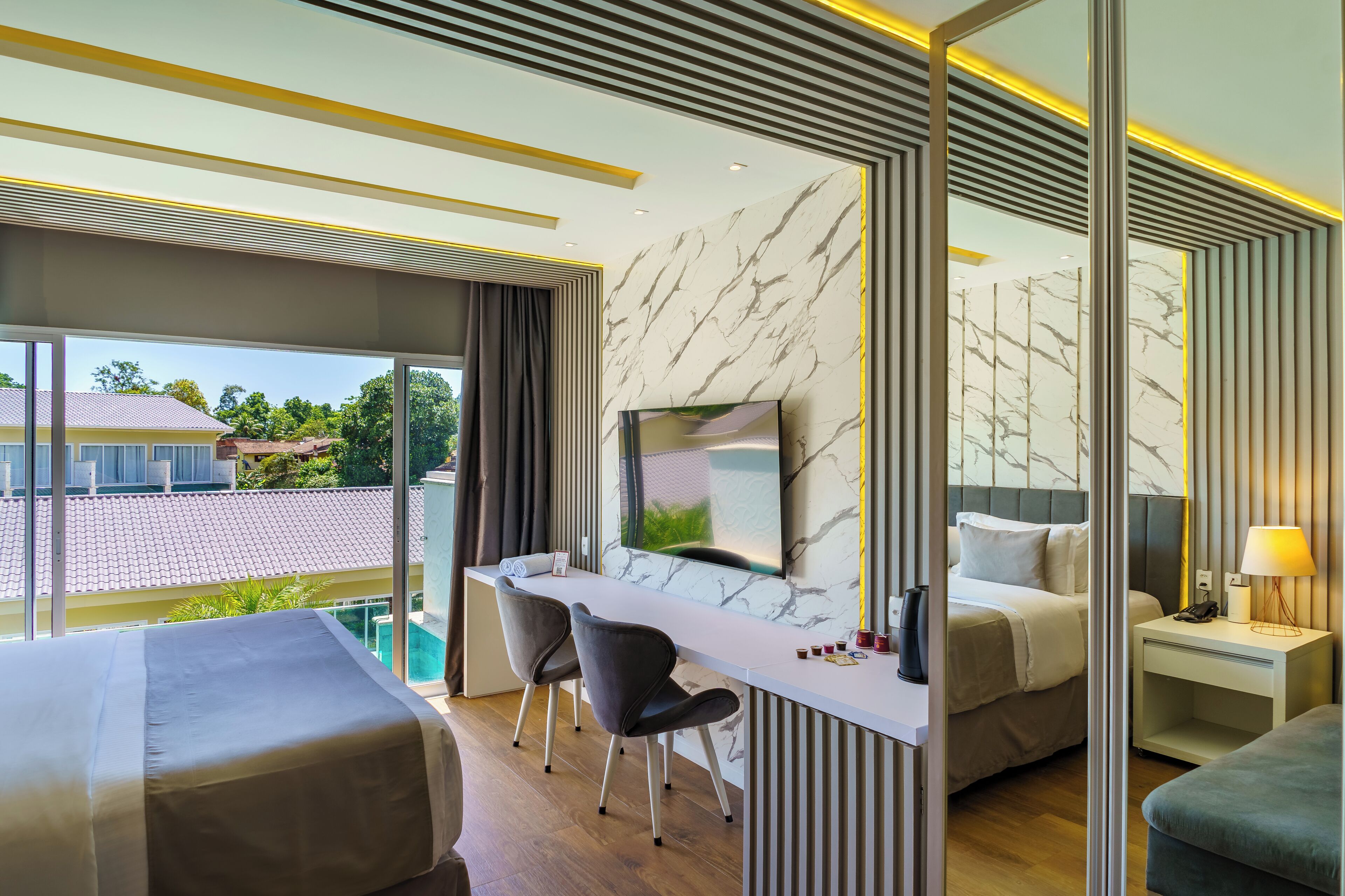 Quarto de hotel moderno com uma cama grande, uma escrivaninha com duas cadeiras, uma televisão e vista para uma piscina e edifícios.