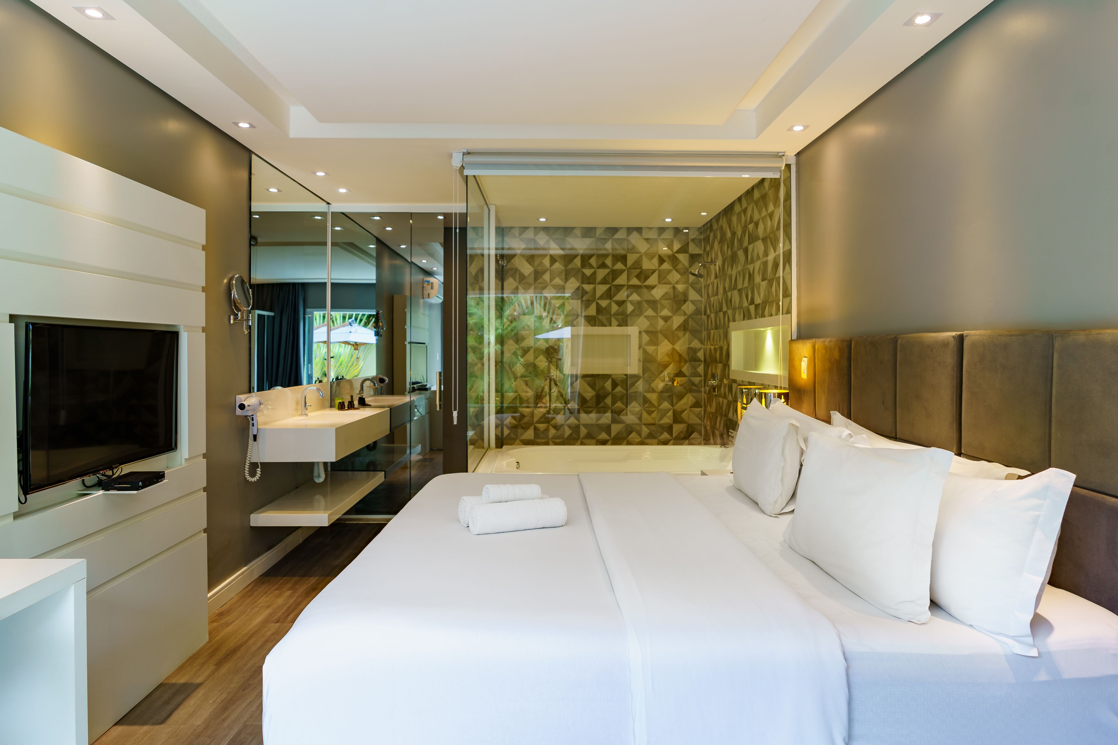 Quarto de hotel moderno com cama grande, televisão, banheiro com chuveiro e parede de vidro.