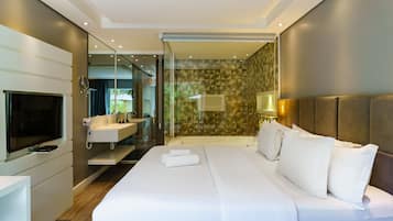 Quarto de hotel moderno com cama grande, televisão, banheiro com chuveiro e parede de vidro.