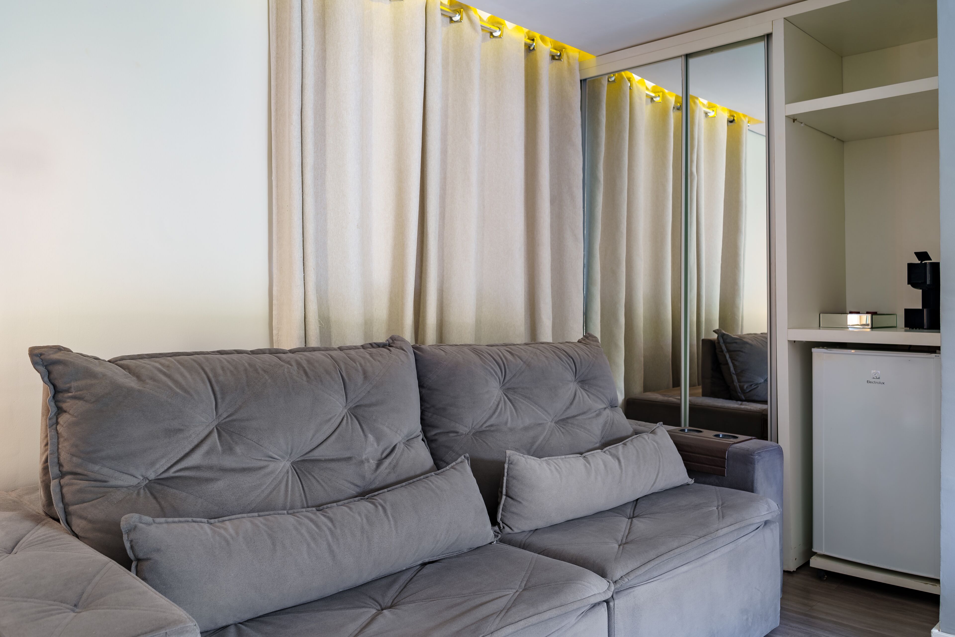 Un sofá gris con varios cojines, un armario blanco y un espejo que refleja la habitación.