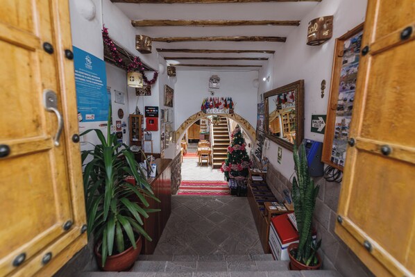 Um corredor estreito com portas de madeira, plantas em vasos, uma árvore de Natal e uma escada que leva a um quarto com cama e mesa de cabeceira.