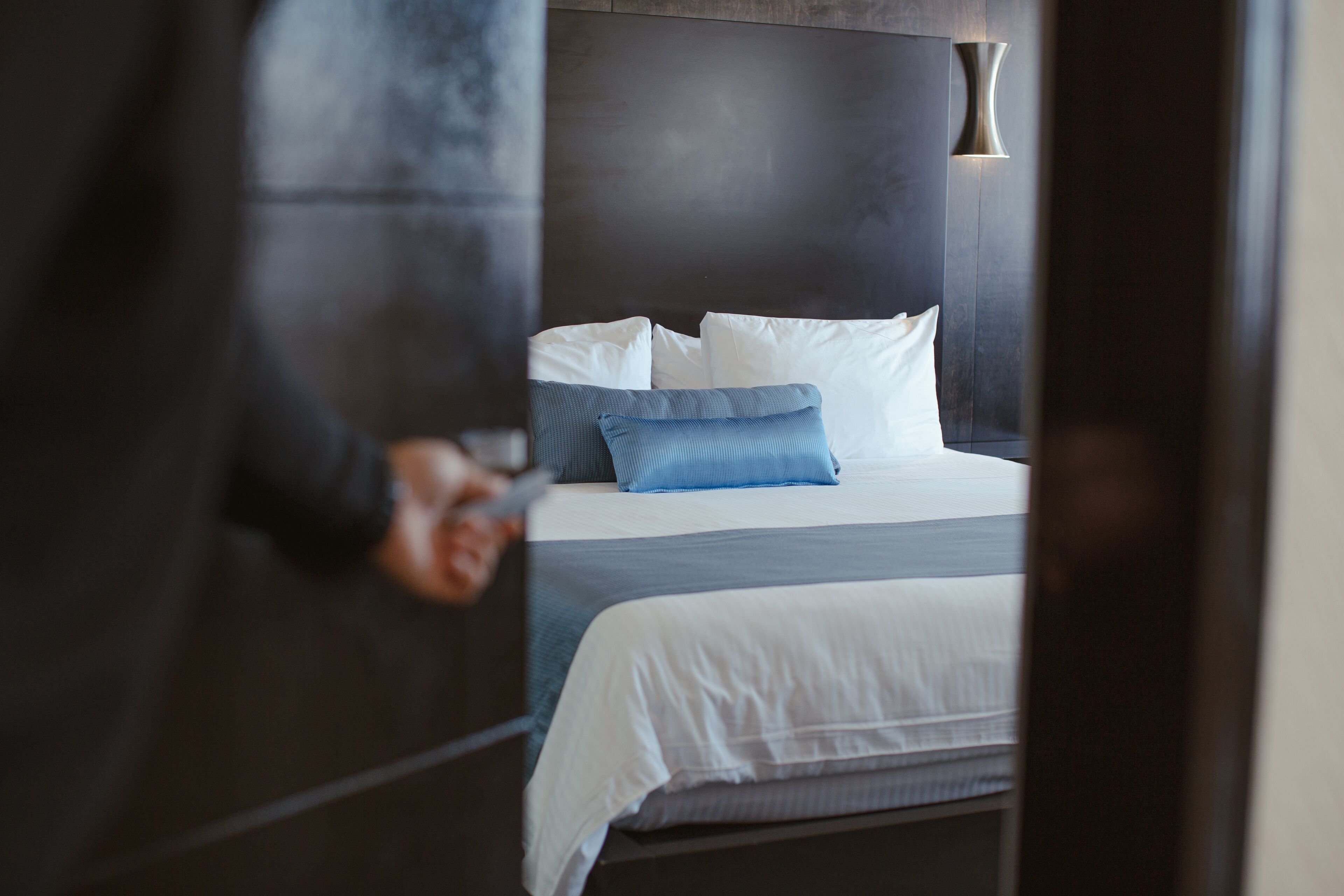 A modern hotel room with a dark wood headboard, a large bed with white and blue bedding, and a wall-mounted light fixture.