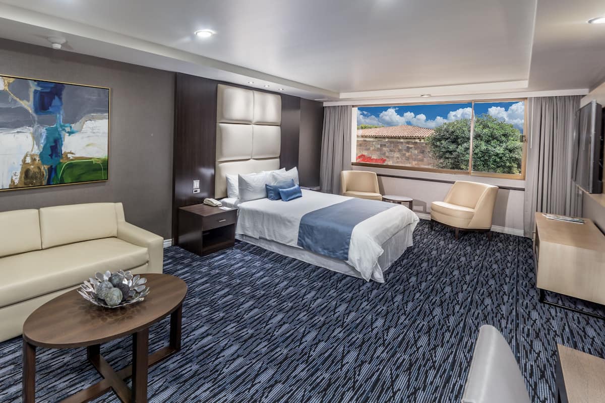 A hotel room with a bed, a sofa, a small table with a decorative bowl, a TV, and a window with a view of a building and trees.