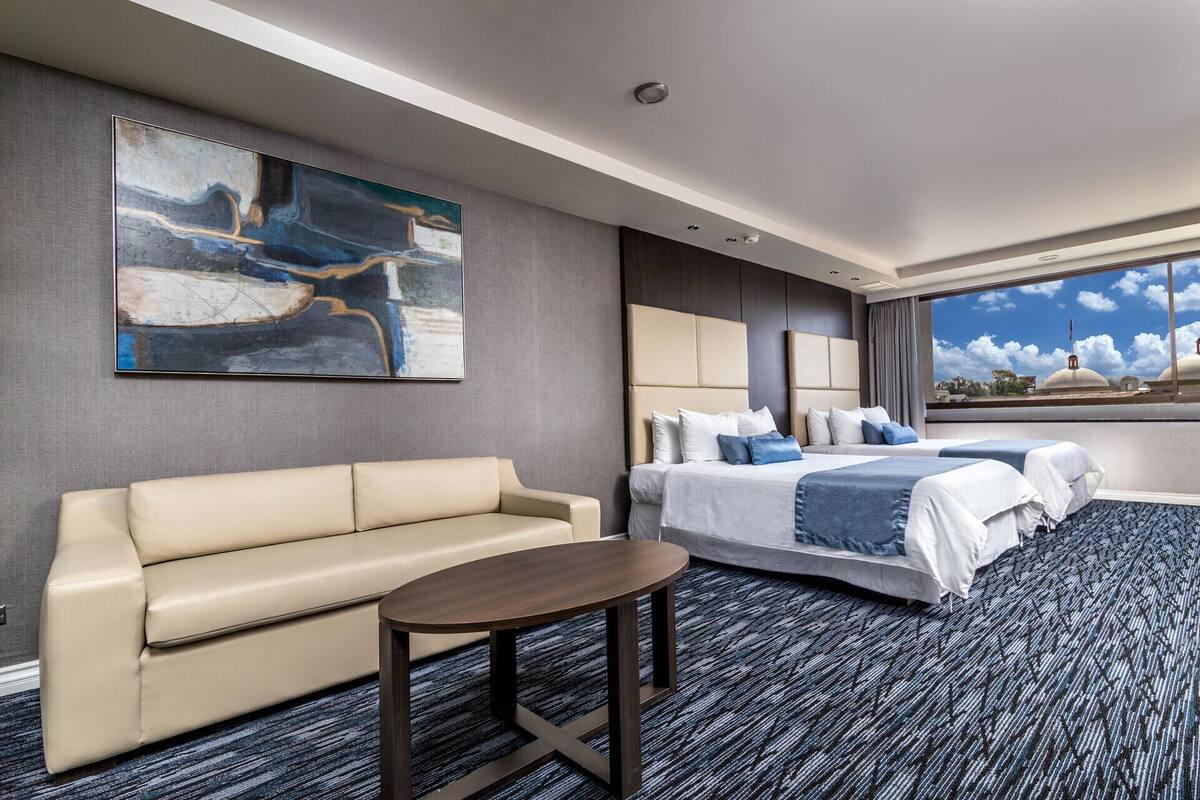 A hotel room with a sofa, a bed with multiple pillows, a small table, and a large abstract painting on the wall.