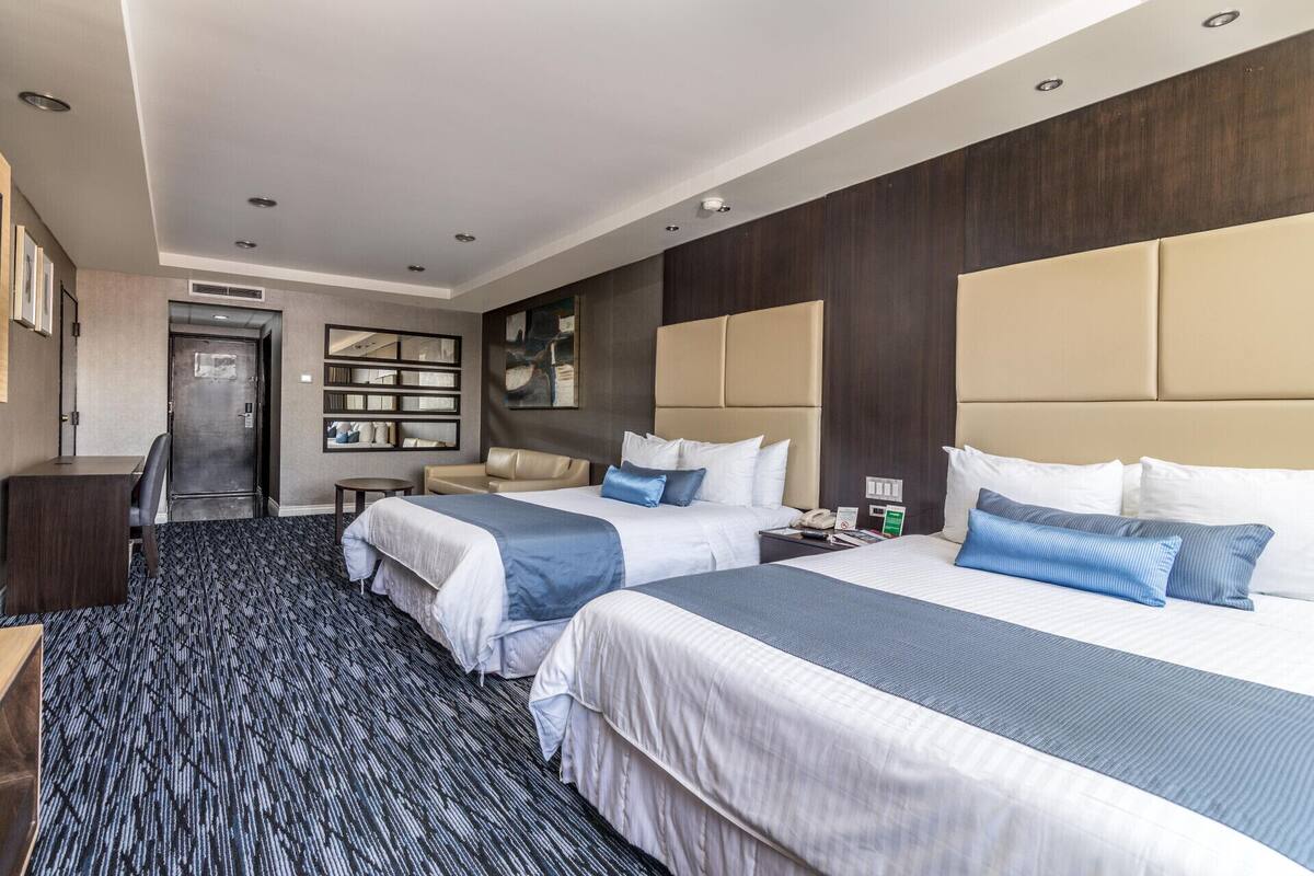 A hotel room with two beds, a desk, a chair, a bookshelf, and a door.