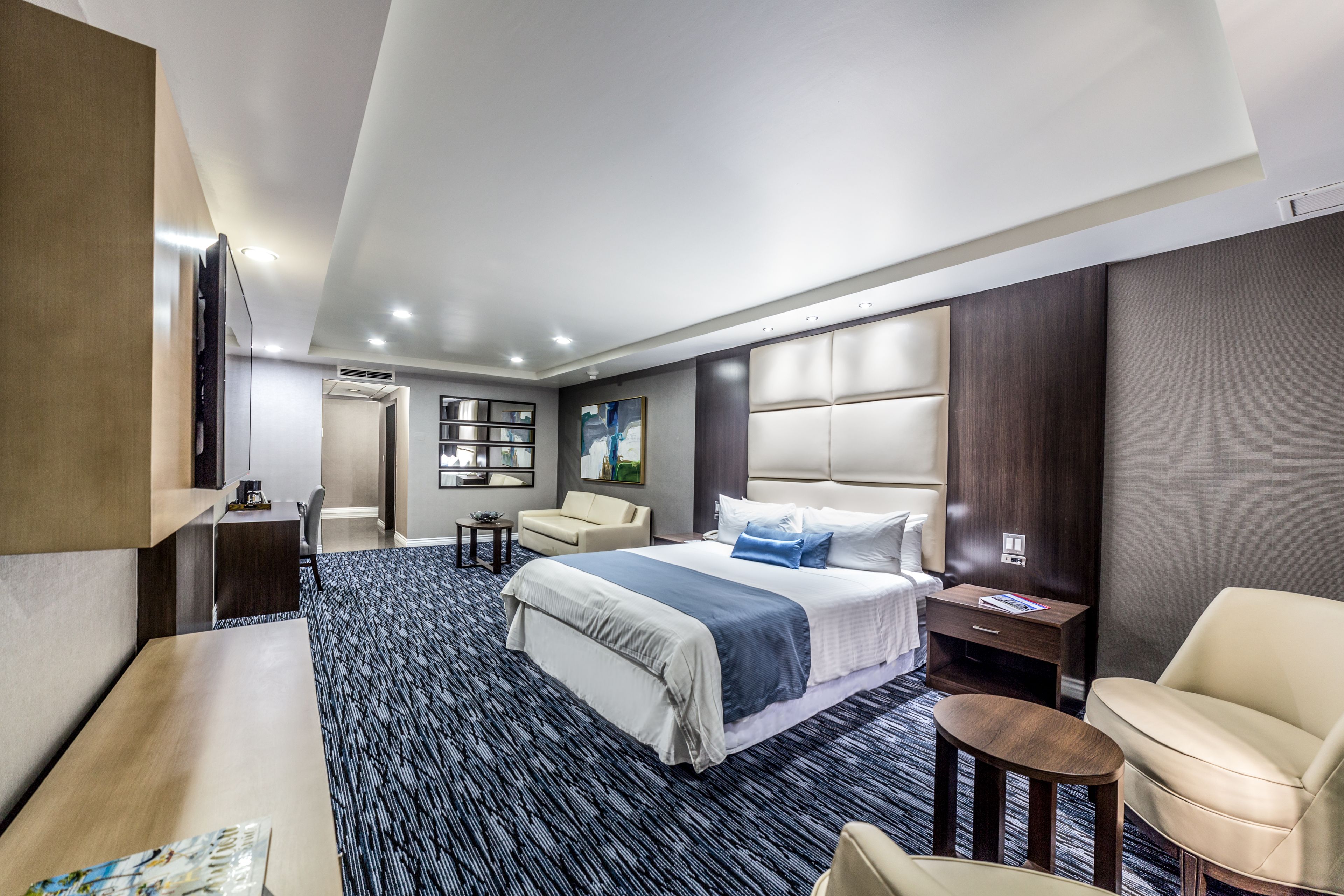 A modern hotel room with a large bed, a desk, a chair, a small table, and a TV.