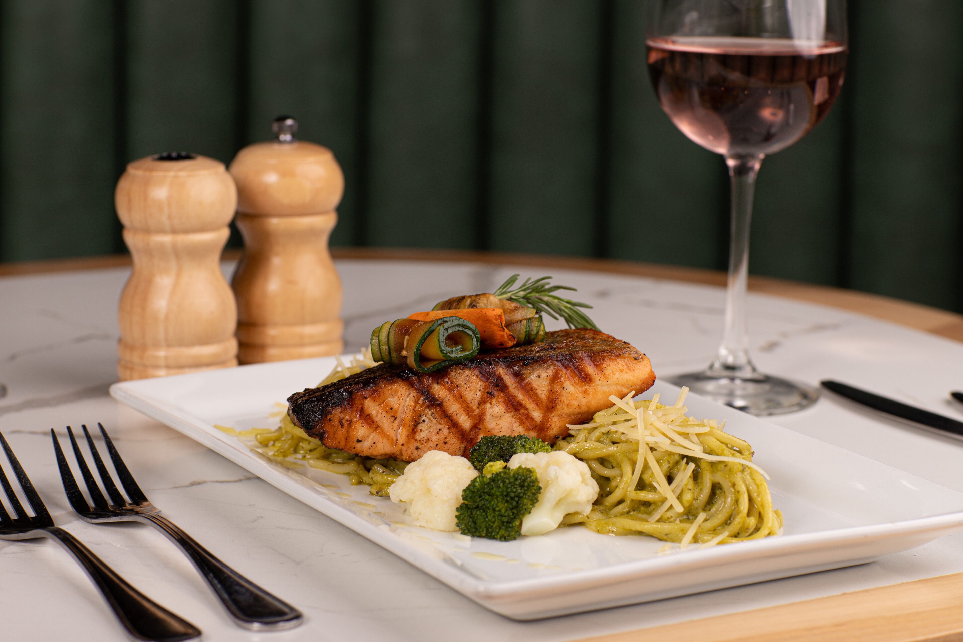 Un plato de comida con salmón, pasta, brócoli y coliflor, acompañado de una copa de vino y saleros y pimenteros sobre una mesa.