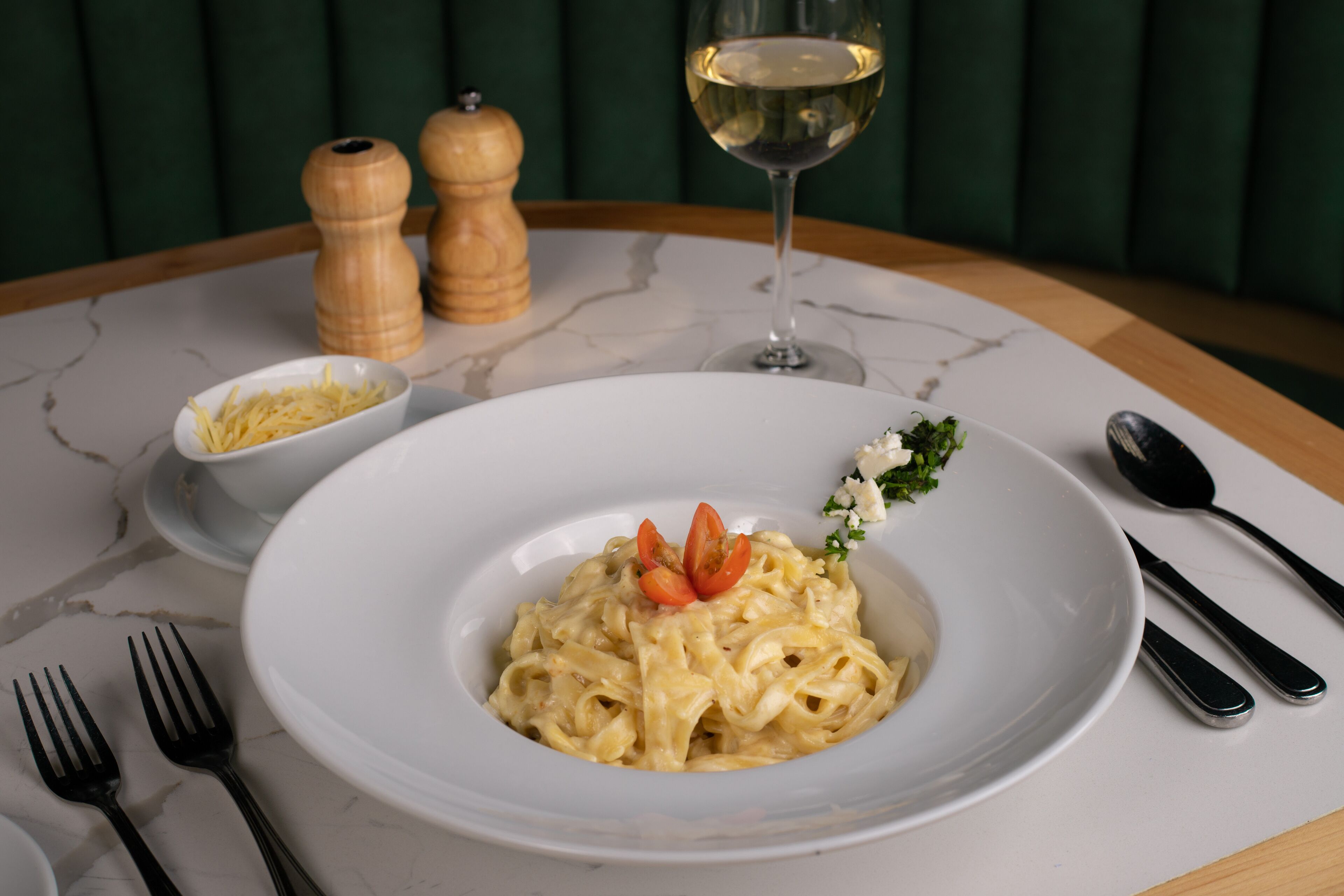 Una mesa de comedor con un plato de pasta, una copa de vino blanco, saleros y pimenteros, y cubiertos.